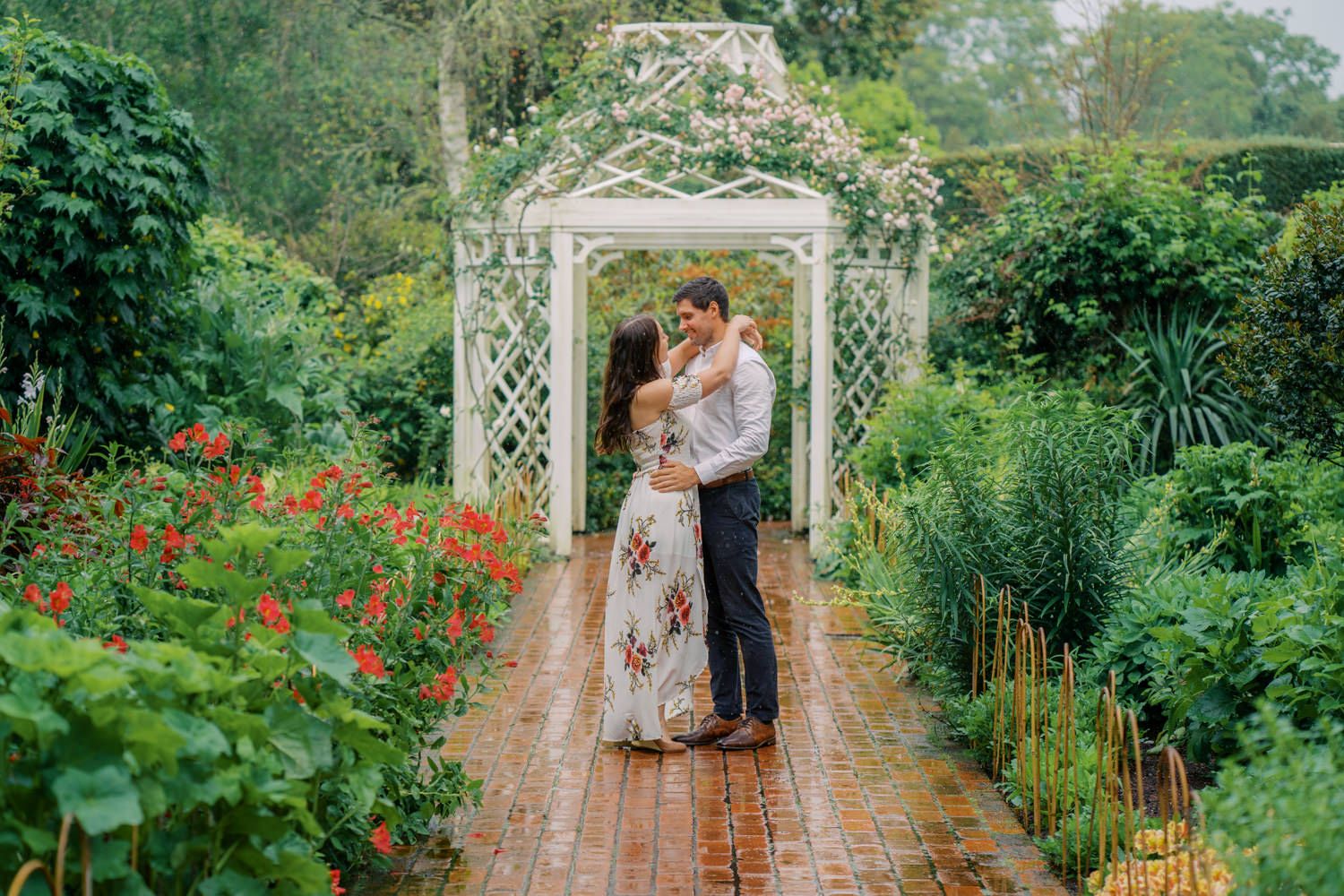 Couple embracing under white garden gazebo surrounded by lush greenery and red flowers on a brick pathway.