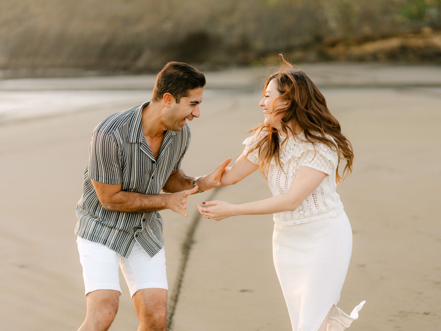 A joyful couple laughs and holds hands while dancing on a sandy beach, dressed in casual white and striped outfits.