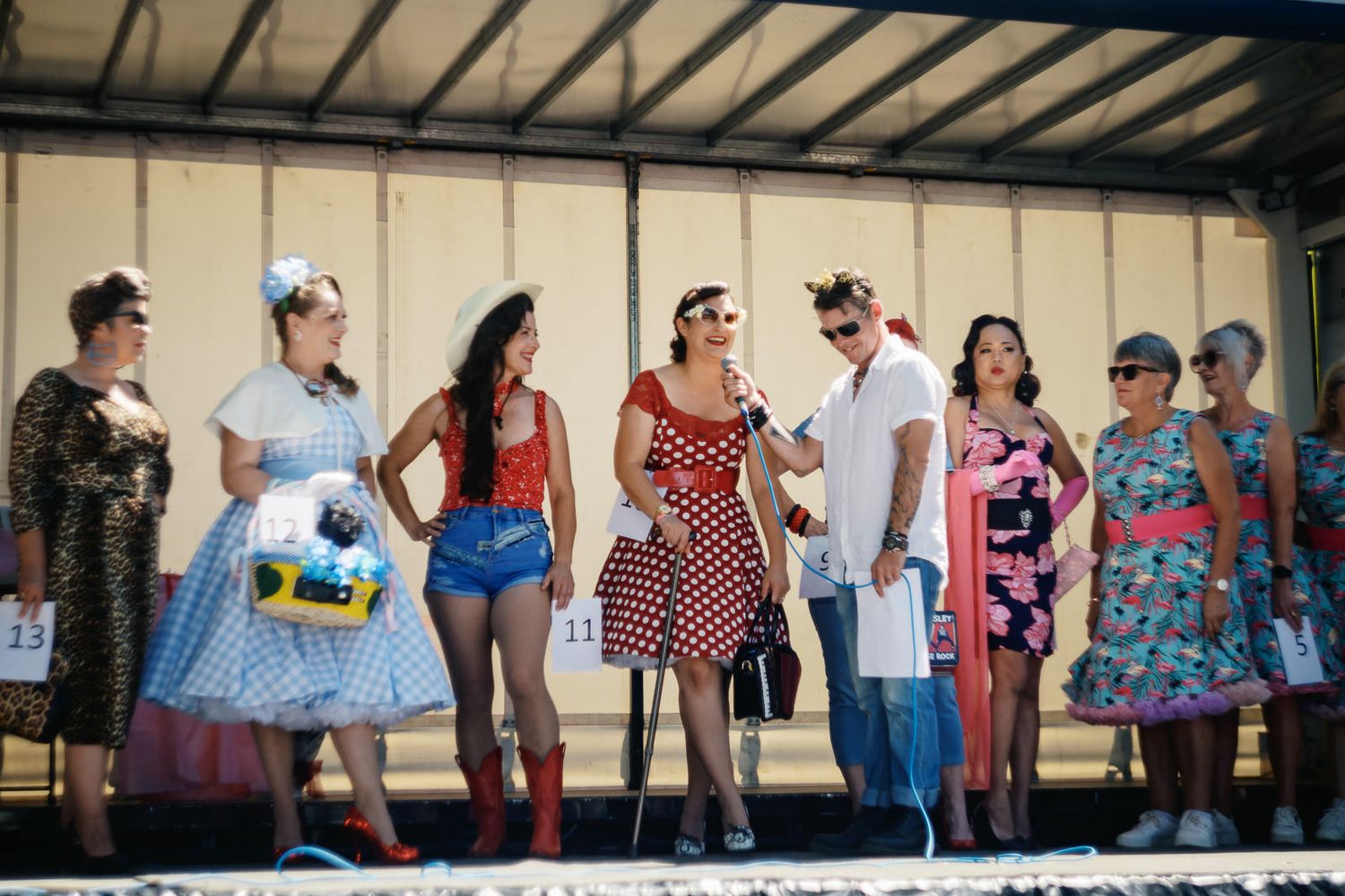 Women in vintage 1950s costumes stand on stage during a retro fashion contest at an outdoor event.
