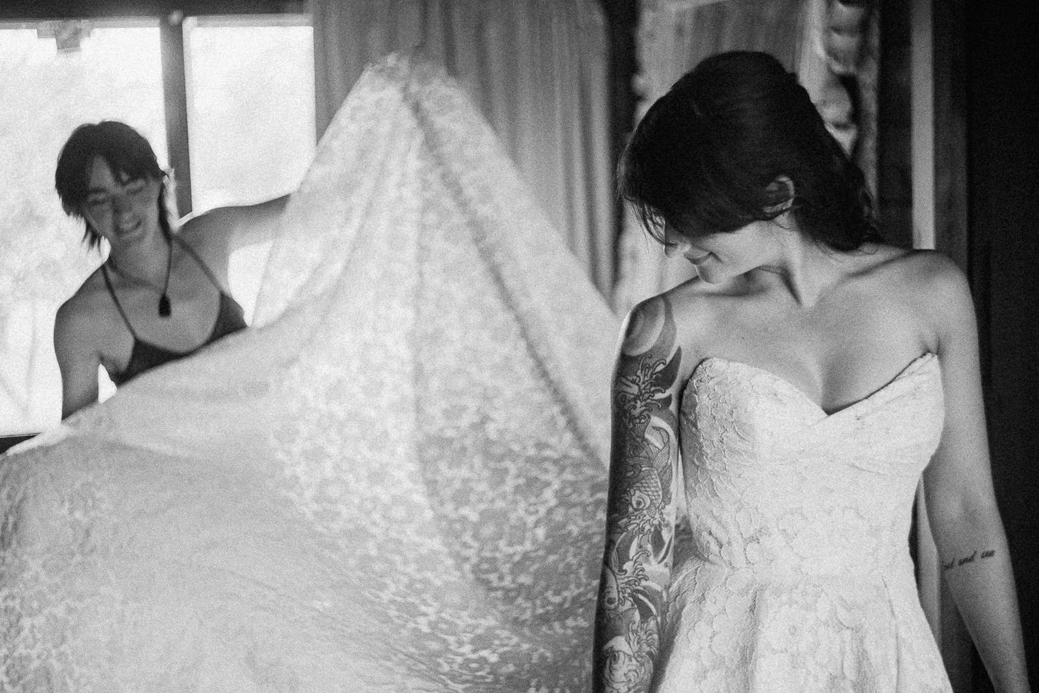 Black and white photo of bride in lace wedding dress with tattooed arm as bridesmaid fluffs veil behind her.