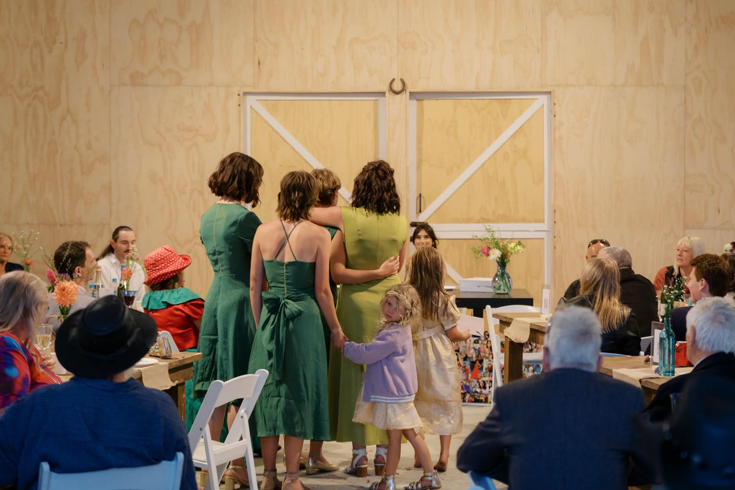 Women in green dresses stand together at an indoor wedding reception with guests seated around them.