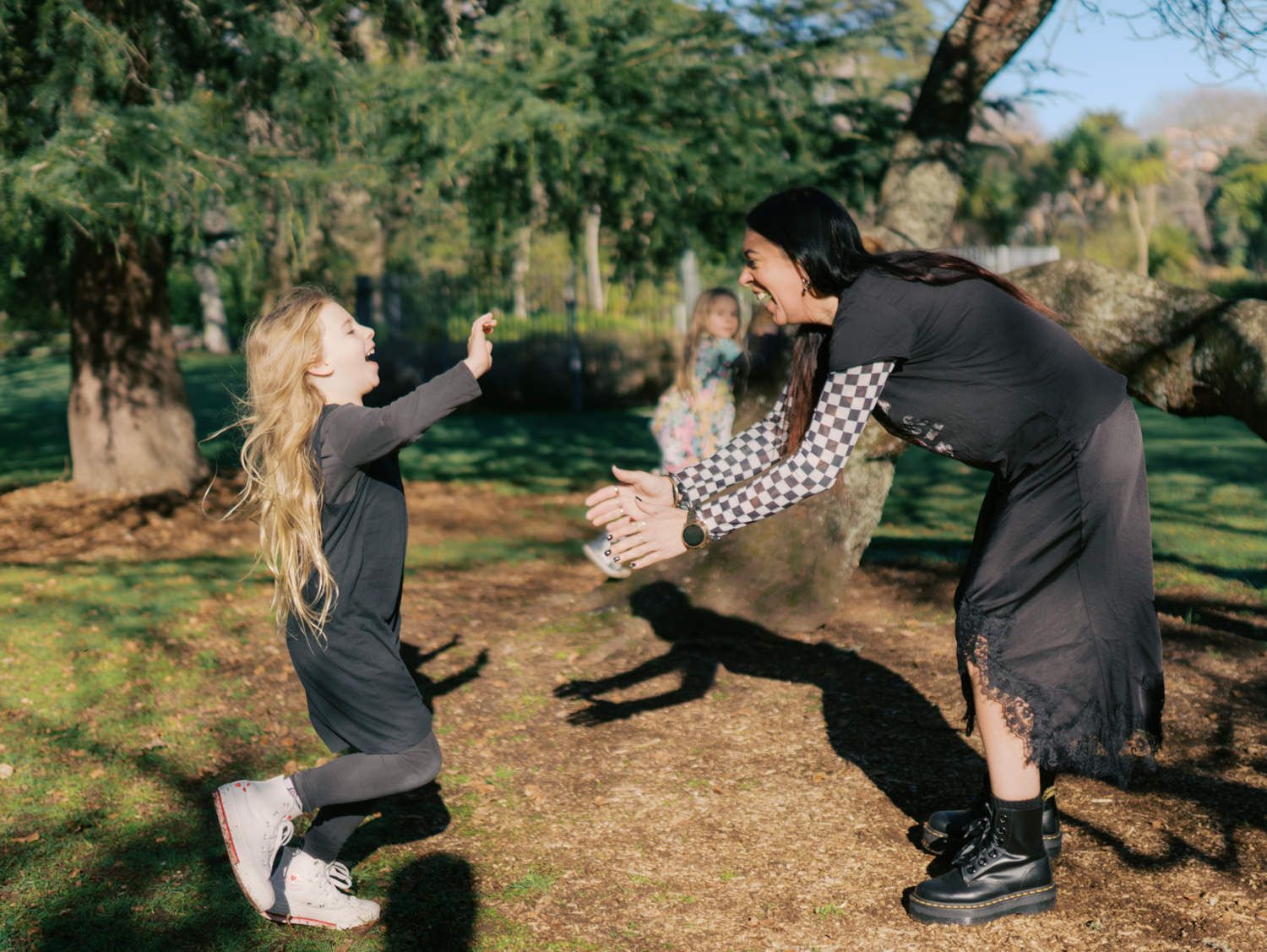 A woman crouching with open arms as a young blonde girl runs toward her in a sunny park.