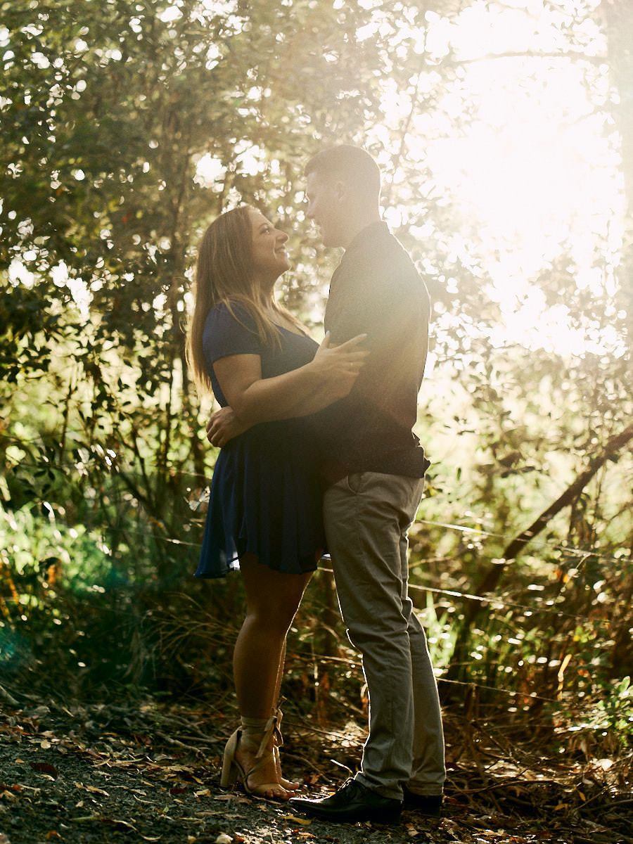 Couple embracing in sunlit forest, woman in blue dress looking up at man, golden backlight filtering through trees.