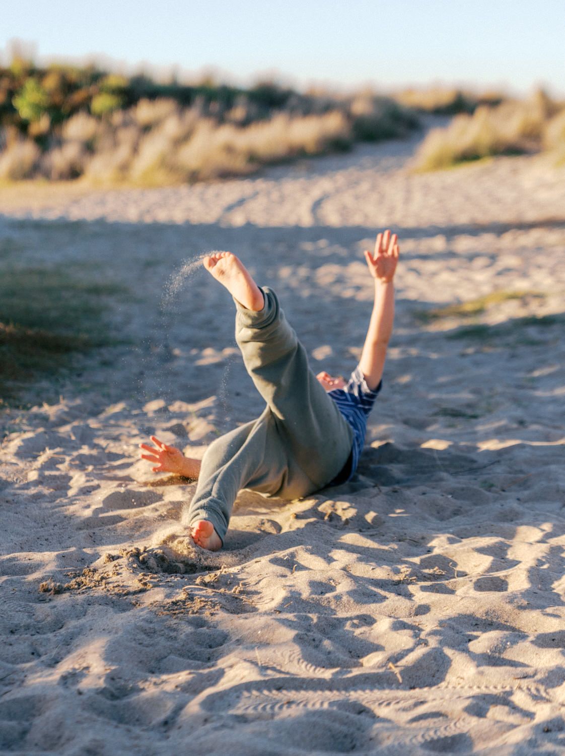 Child playing in sandy beach, falling backwards with arms raised in joy on a sunny day.