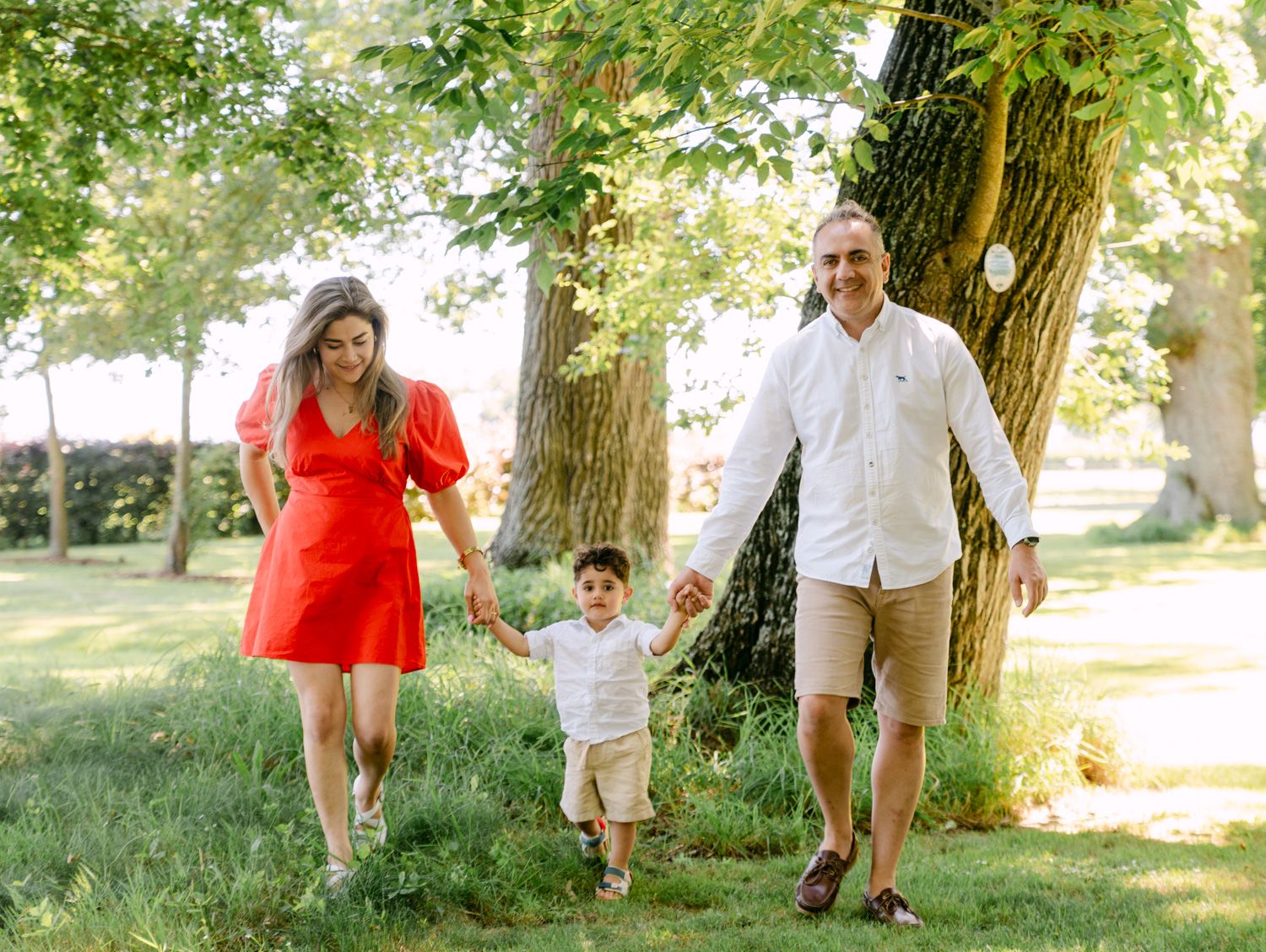 Happy family of three walks hand-in-hand through a sunny green park, mom in red dress and dad in white shirt.