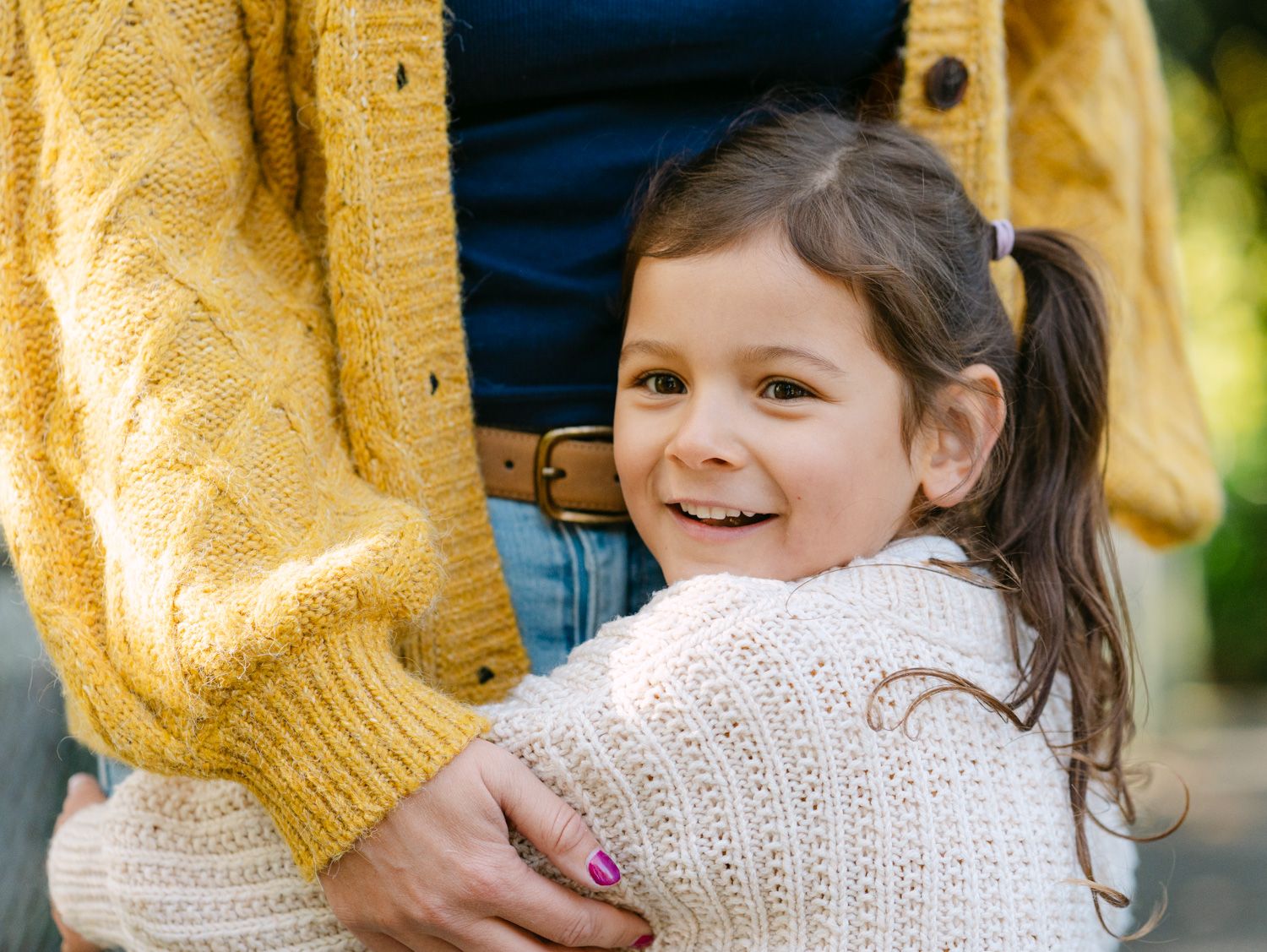 A young girl in a white knit sweater smiles while hugging an adult wearing a yellow sweater outdoors.