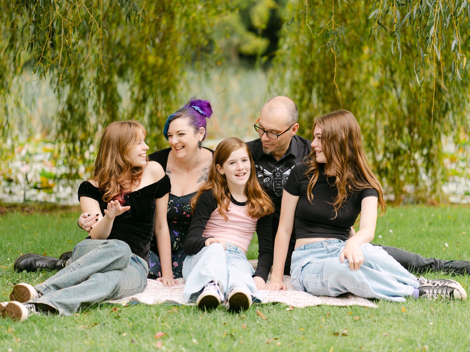 Happy family of five sitting on grass in a lush green park, smiling and enjoying time together outdoors.