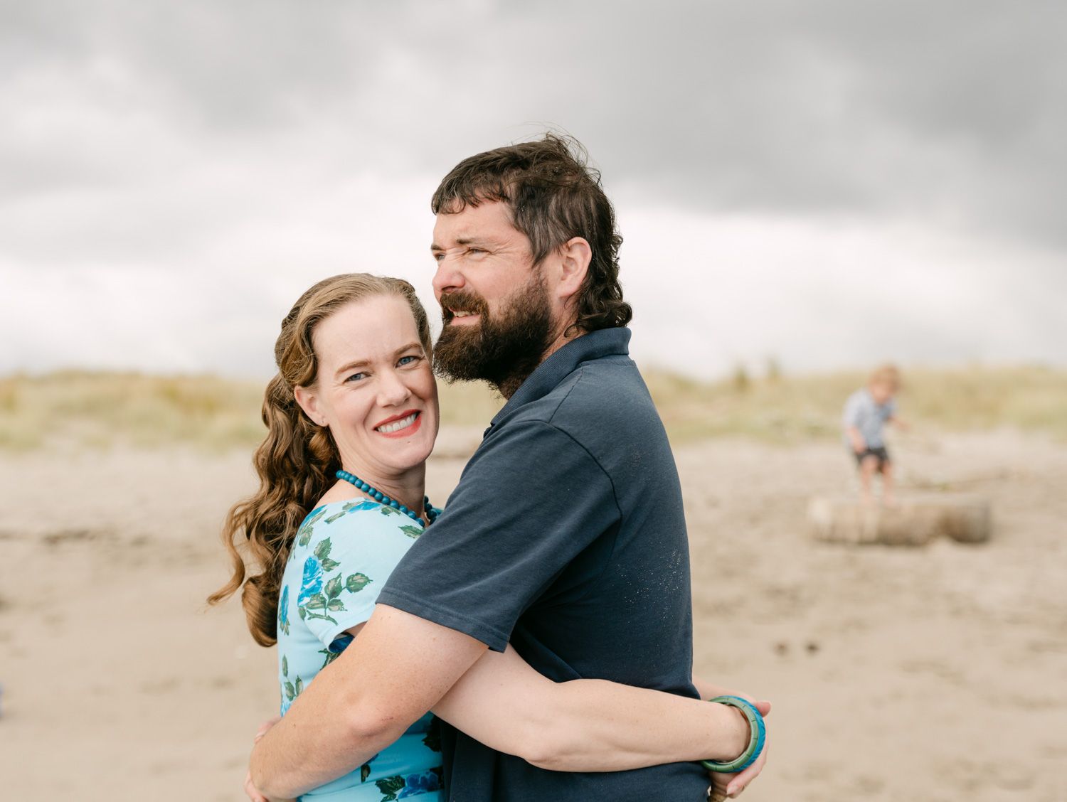 A smiling couple embraces on a sandy beach under cloudy skies, with a child visible in the background.