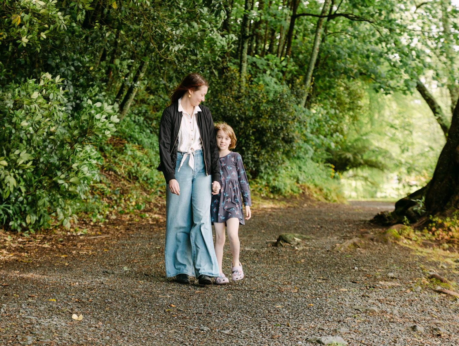 A woman and young girl walk hand-in-hand on a gravel path surrounded by lush green trees.