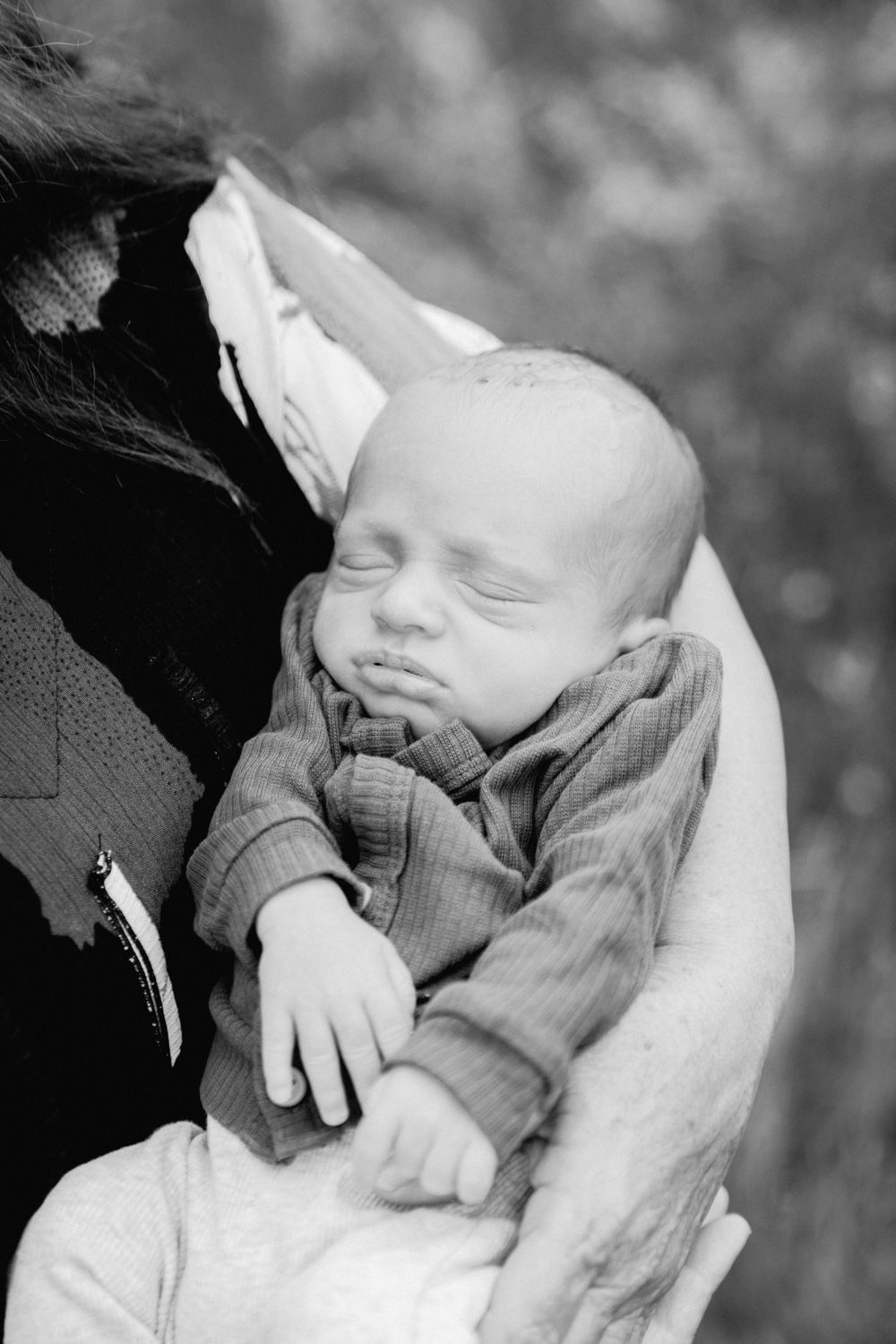 Black and white photo of a sleeping newborn baby cradled in a parent's arms outdoors.