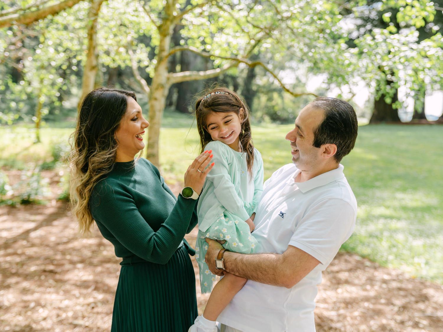 Family portrait of parents and young daughter in a lush green park, sharing a joyful moment together.