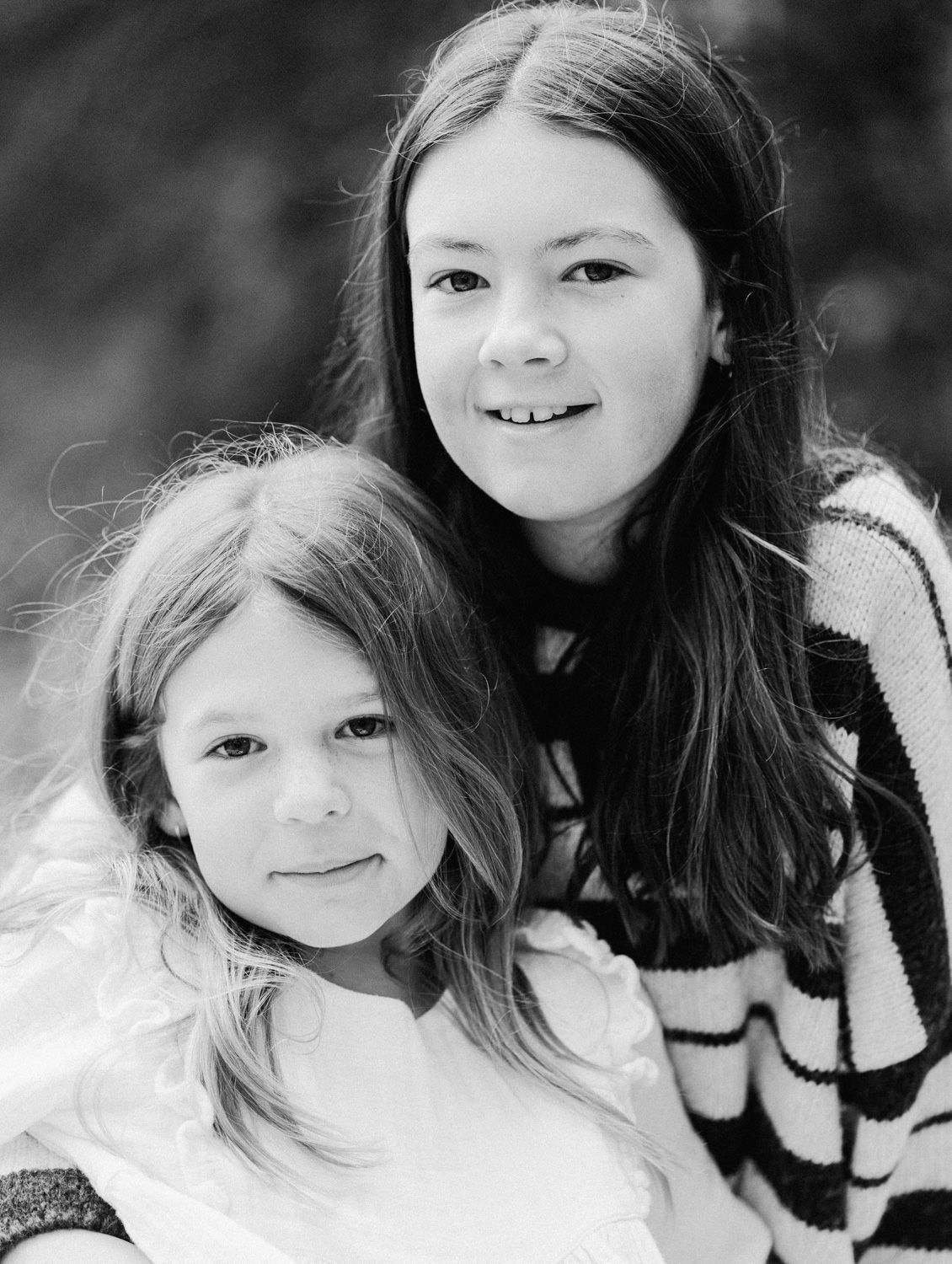 Black and white portrait of two young sisters smiling together outdoors, older girl in striped sweater hugging younger sibling.