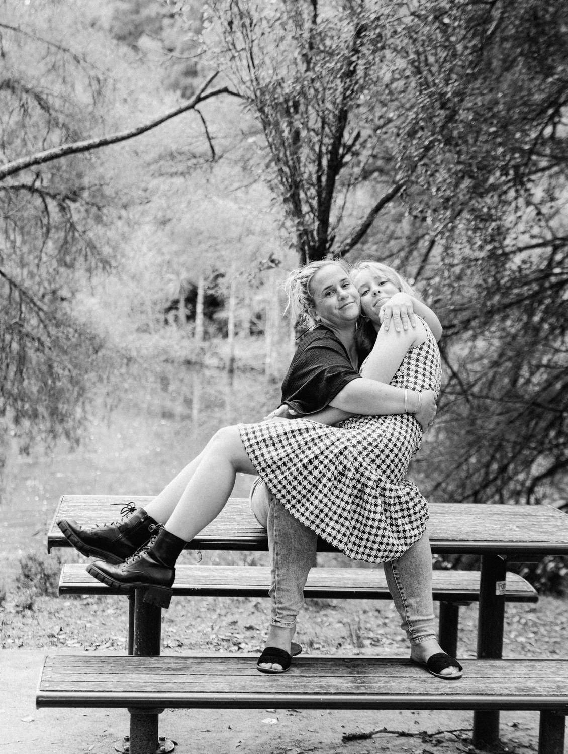 Two young women embrace on a park bench surrounded by trees in a black and white outdoor photo.