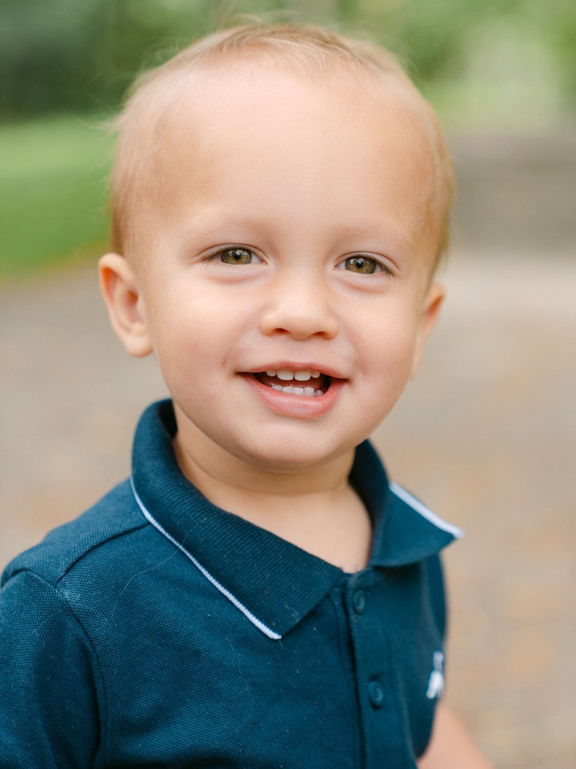 Smiling toddler boy in navy polo shirt posing outdoors in a park setting.