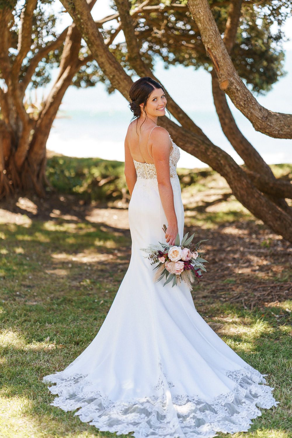Bride in white mermaid lace wedding gown holding floral bouquet, posing under trees near beach.
