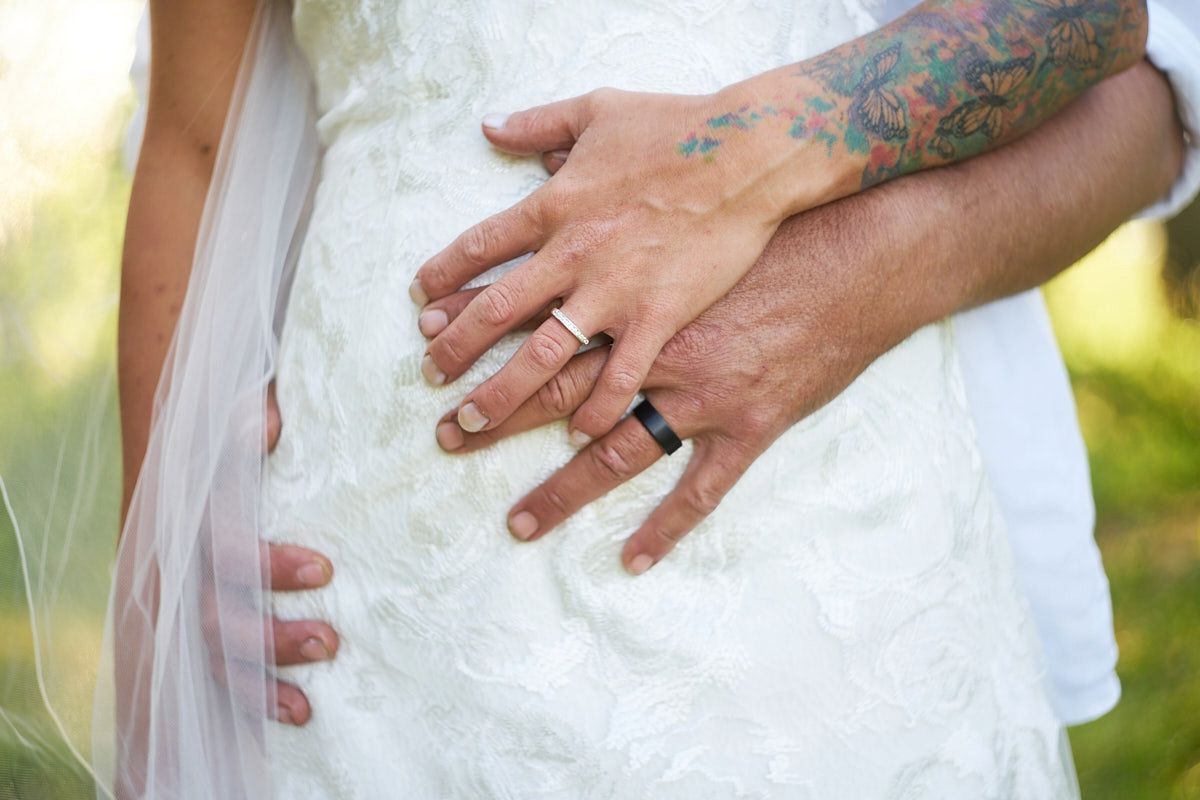 Bride and groom showing wedding rings on hands, groom with tattoo sleeve, touching bride's white lace wedding dress.