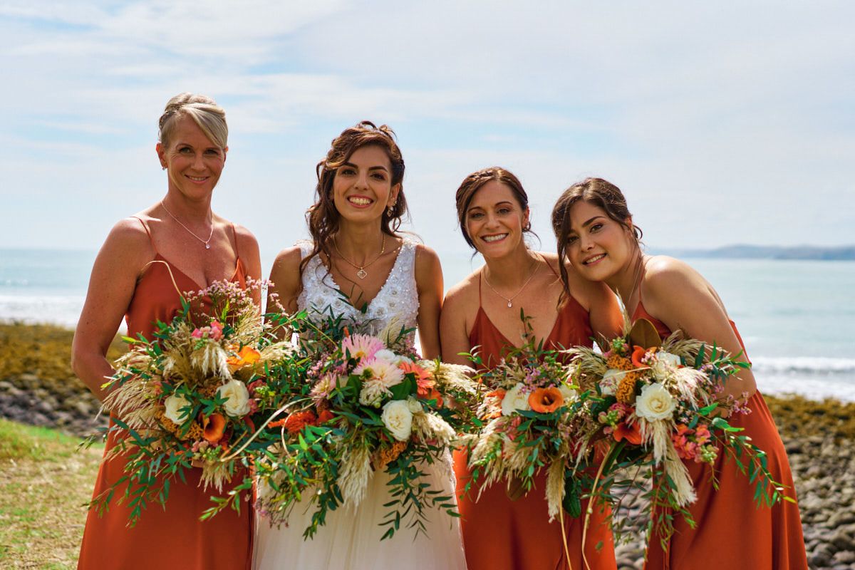 Bride in white gown with three bridesmaids in rust orange dresses holding lush bouquets by the ocean.