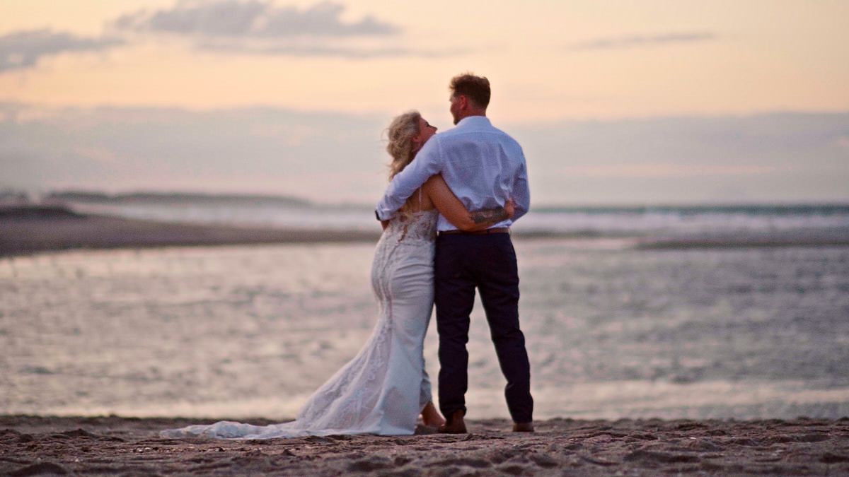 Bride and groom share a romantic embrace while watching the sunset over the ocean on a sandy beach.