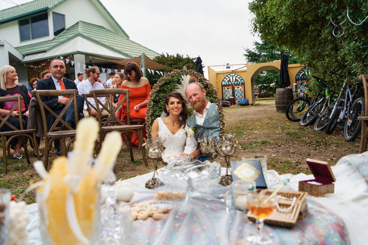 Happy couple smiles at outdoor wedding reception table with food and drinks, guests seated in background.