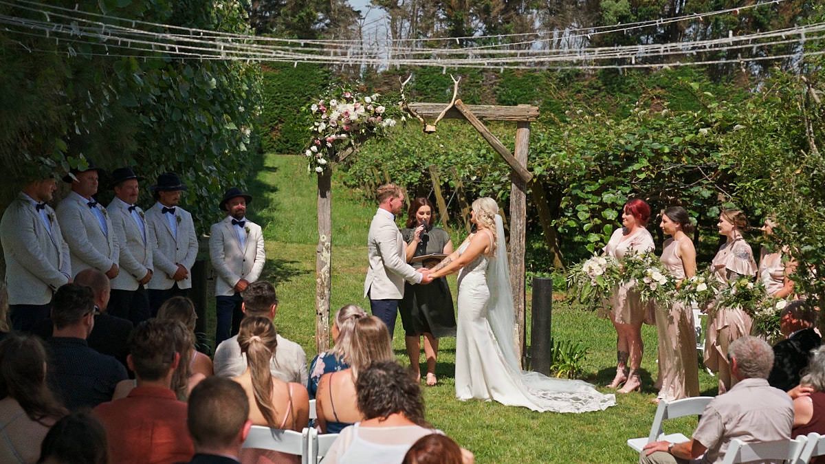 Outdoor wedding ceremony with couple at floral arch, groomsmen in white, bridesmaids with bouquets, and guests seated on sunny lawn.