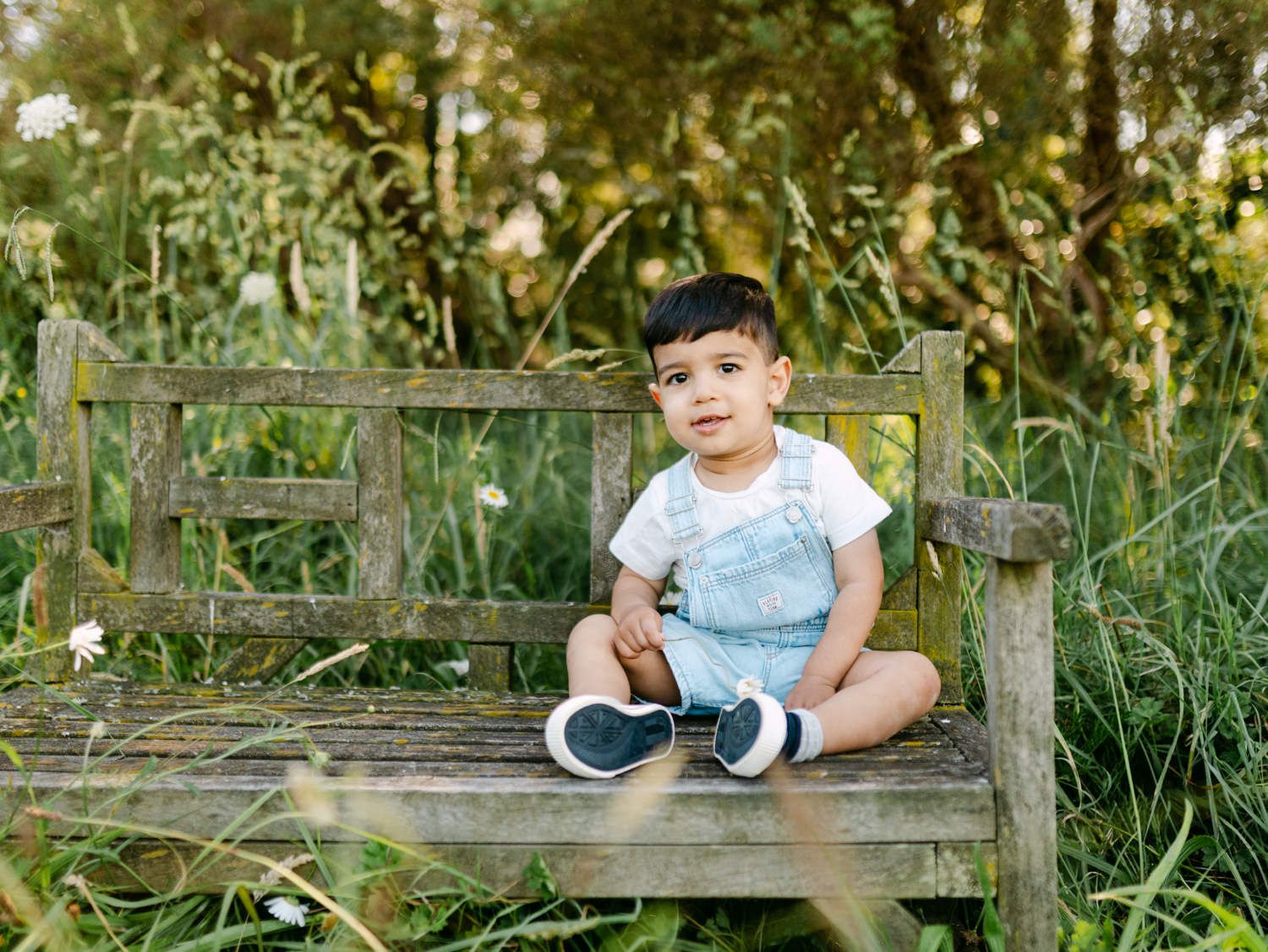 A toddler boy in denim overalls sits on a rustic wooden bench surrounded by lush green garden foliage.