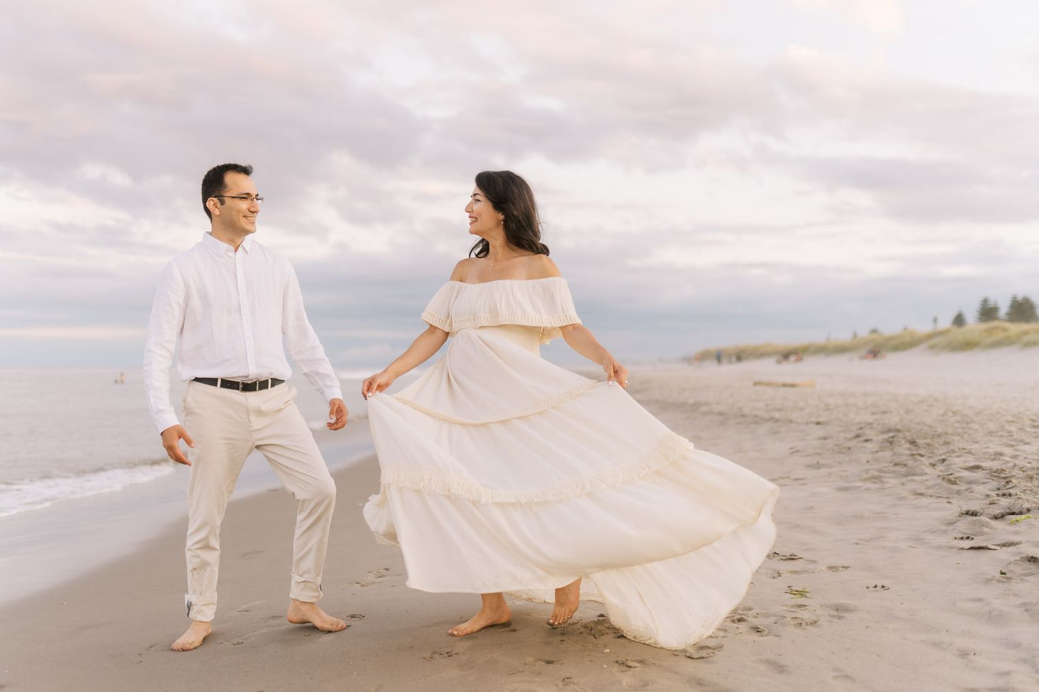 Couple in white outfits walking hand in hand on a beach, woman's flowing dress blowing in the wind.