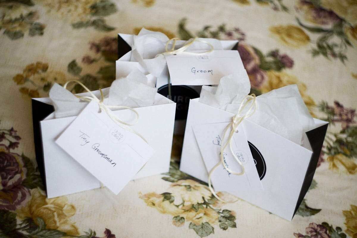 White wedding gift bags with twine ties and tags labeled Groom displayed on a floral tablecloth.