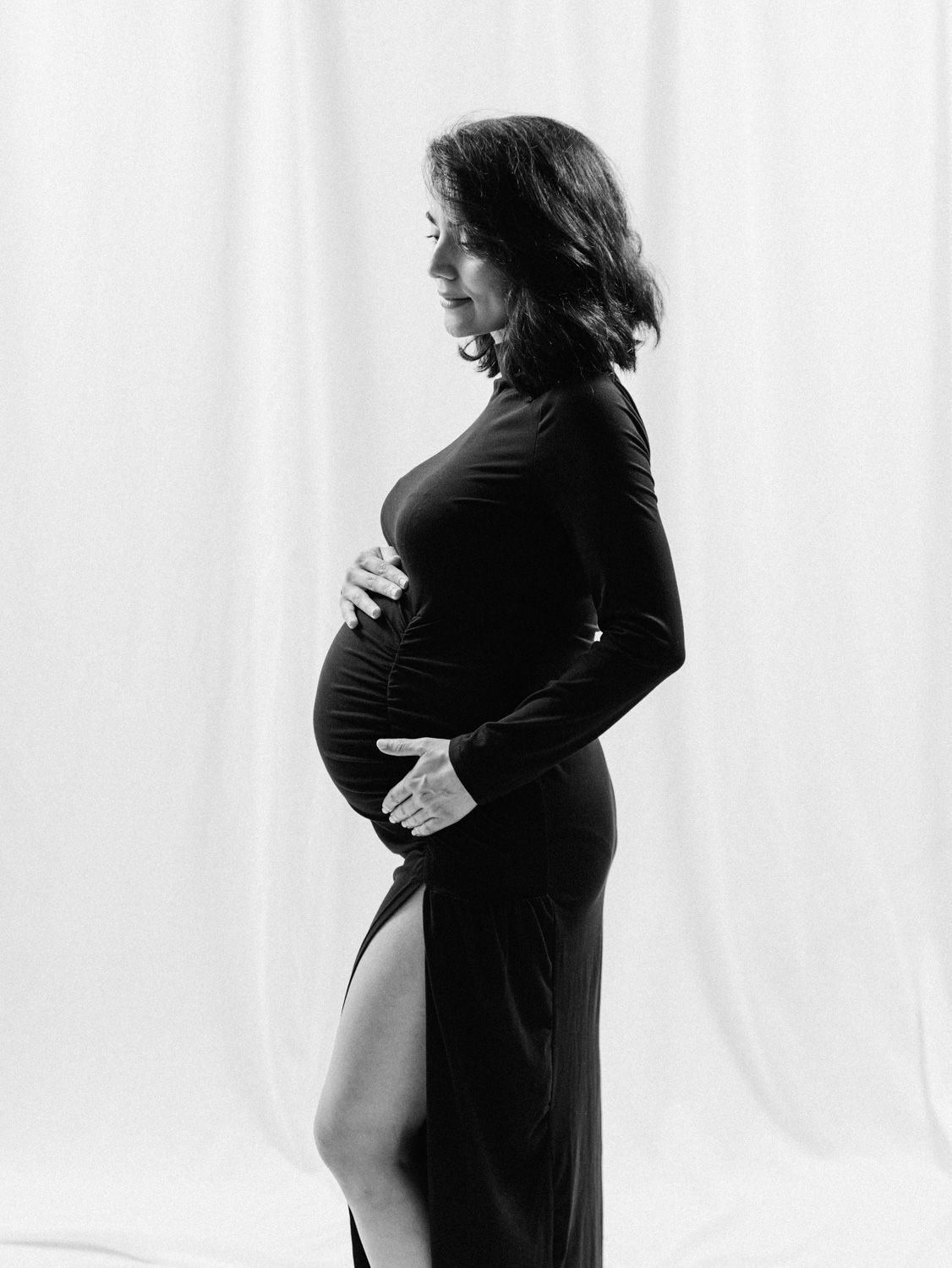 Elegant black and white side profile of pregnant woman in black gown holding baby bump in studio.