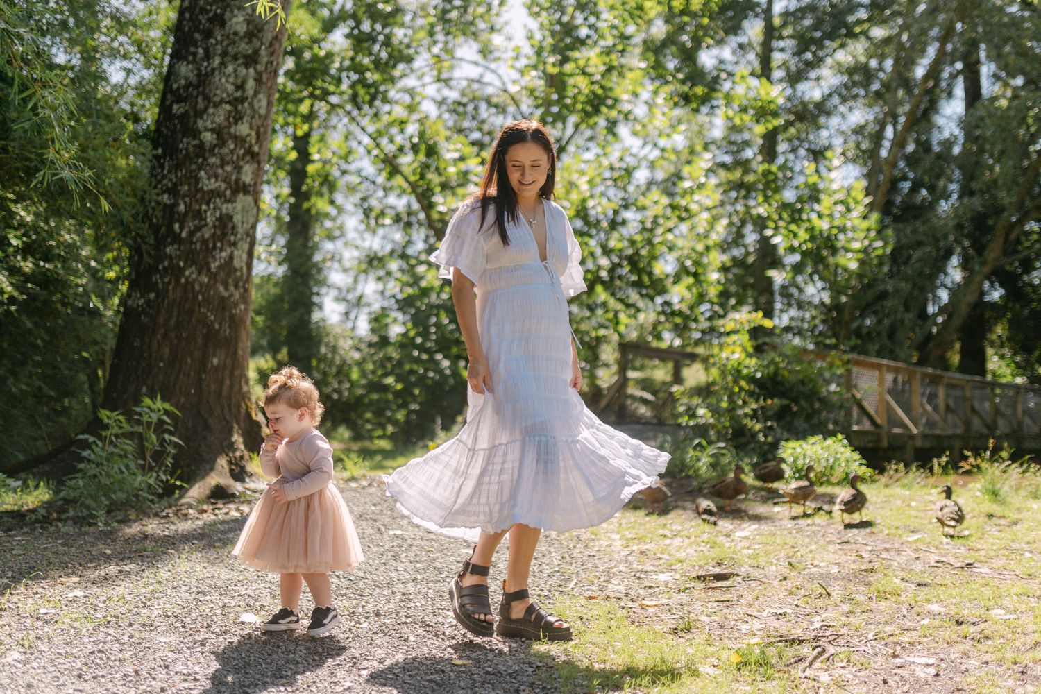 Pregnant woman in white dress twirling with toddler girl in pink dress on sunny gravel path surrounded by trees.