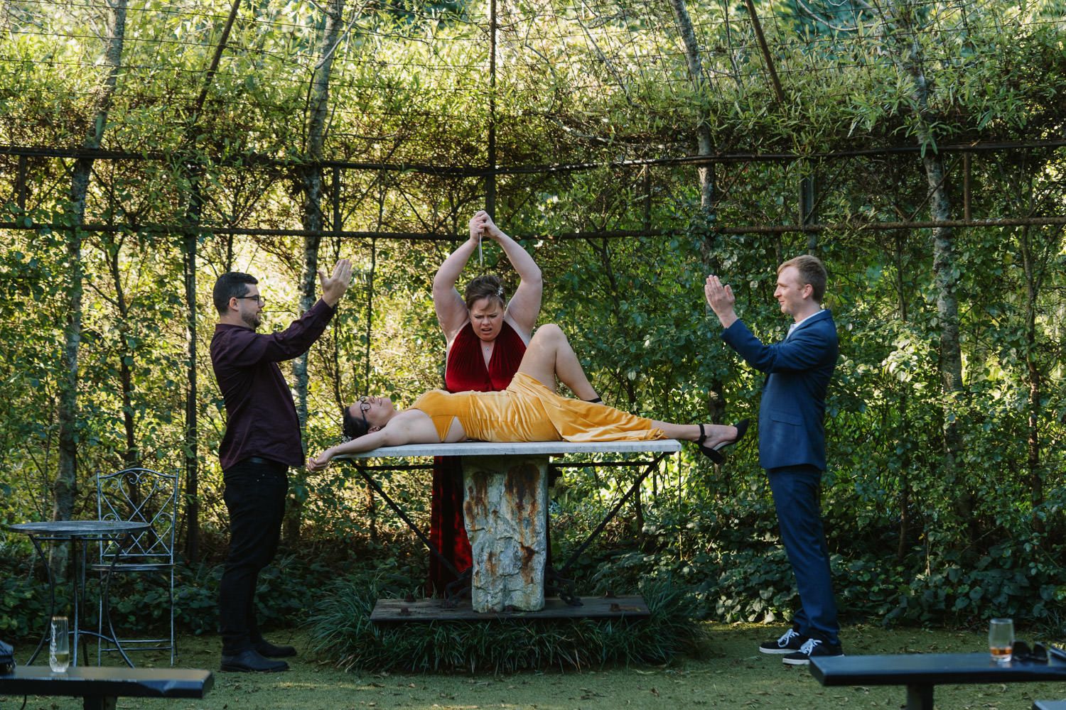 Three performers in colorful outfits doing an acrobatic balancing act outdoors in a lush green garden setting.