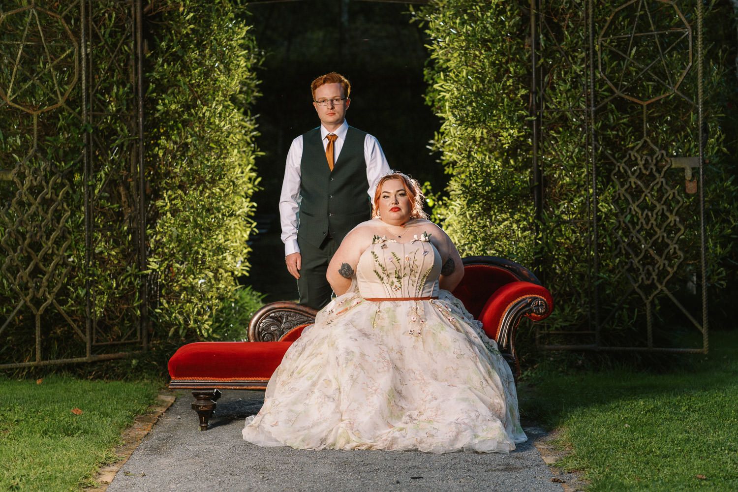 Couple poses in garden wedding photo, groom standing behind bride in ornate white ballgown on red velvet chaise.