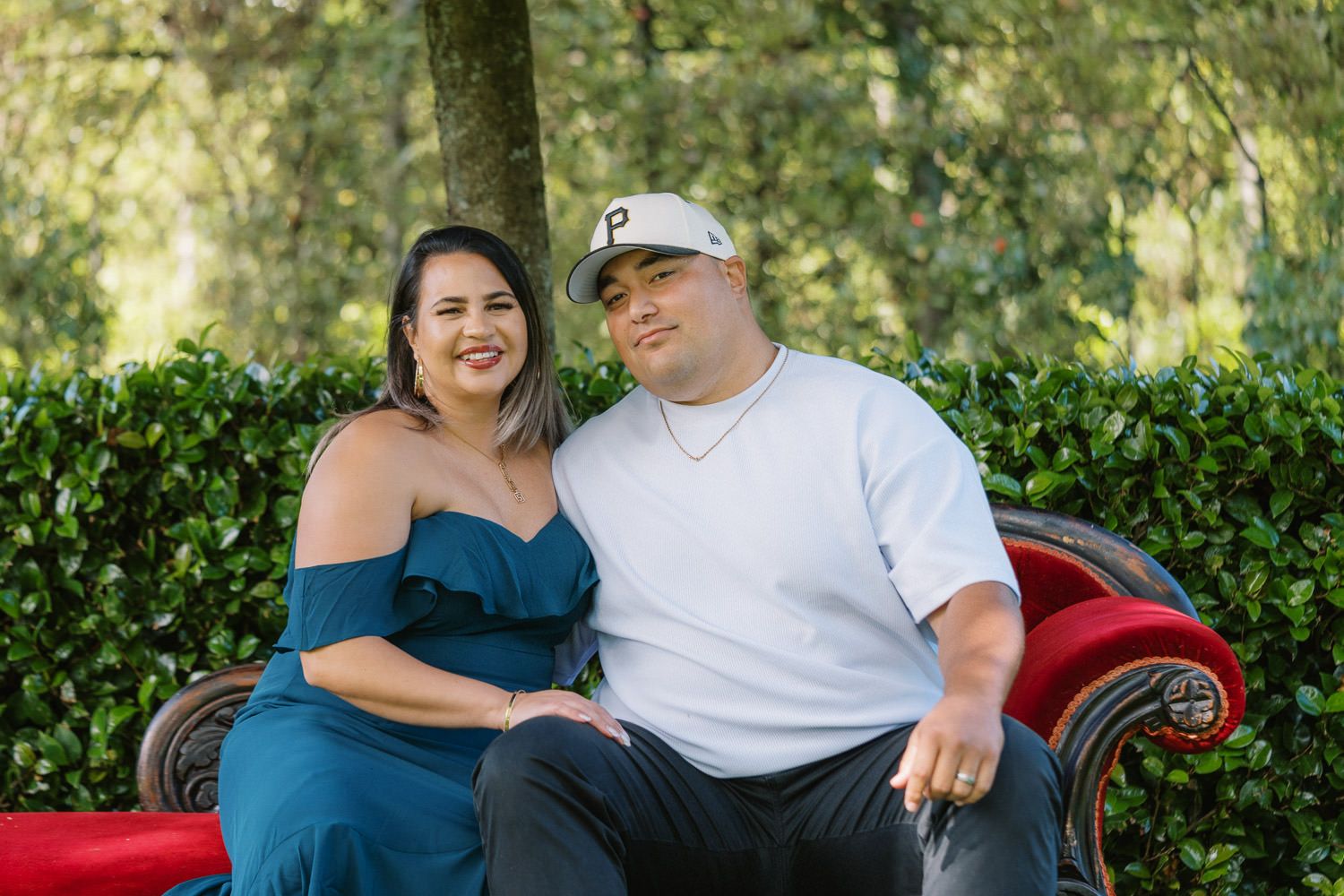 Couple sitting on a red velvet bench in a garden, woman in blue dress and man in white shirt and cap.