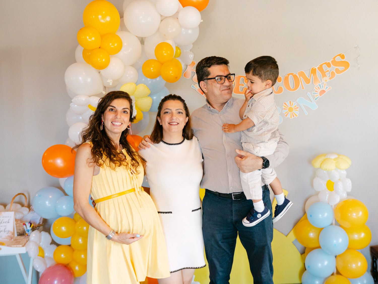 Family posing at a baby shower with colorful balloons, pregnant woman in yellow dress, man holding toddler.