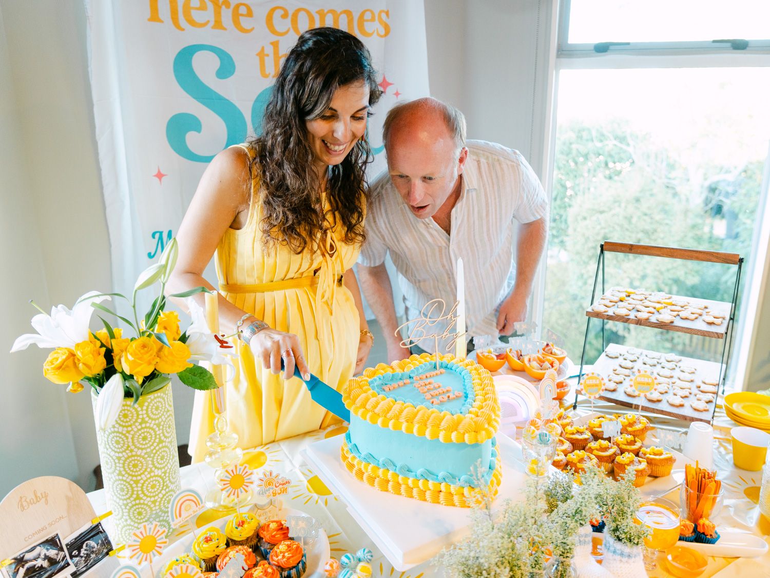 Couple cutting a blue heart-shaped baby shower cake at a festive table with yellow floral decor.