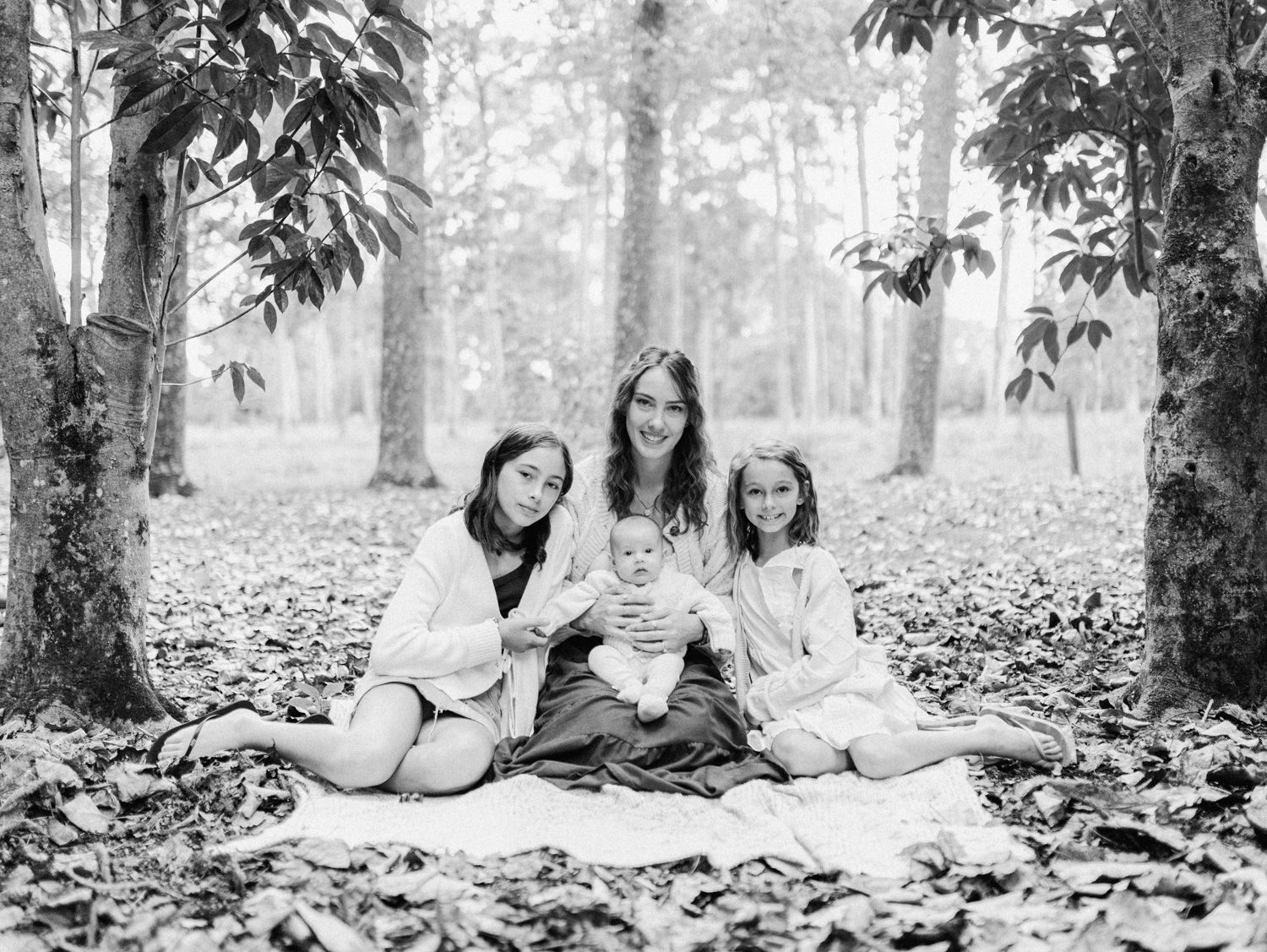 Black and white photo of a mother and three children sitting on a blanket among fallen leaves in a forest.