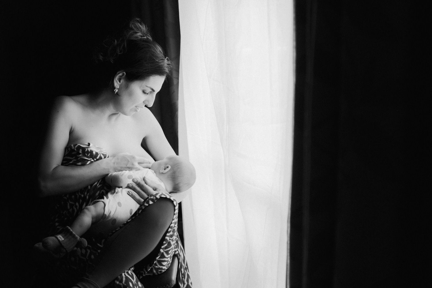 Black and white photo of a mother breastfeeding her newborn baby by a sunlit window.
