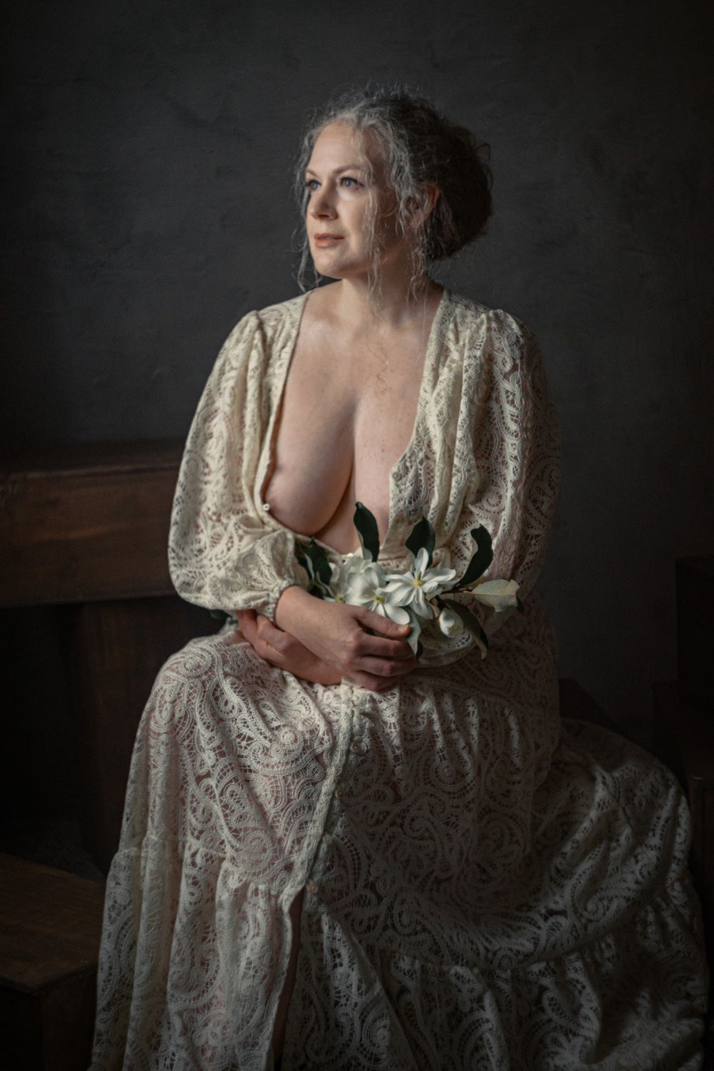 Elegant older woman in lace dress holding white flowers, seated against dark background in artistic portrait.