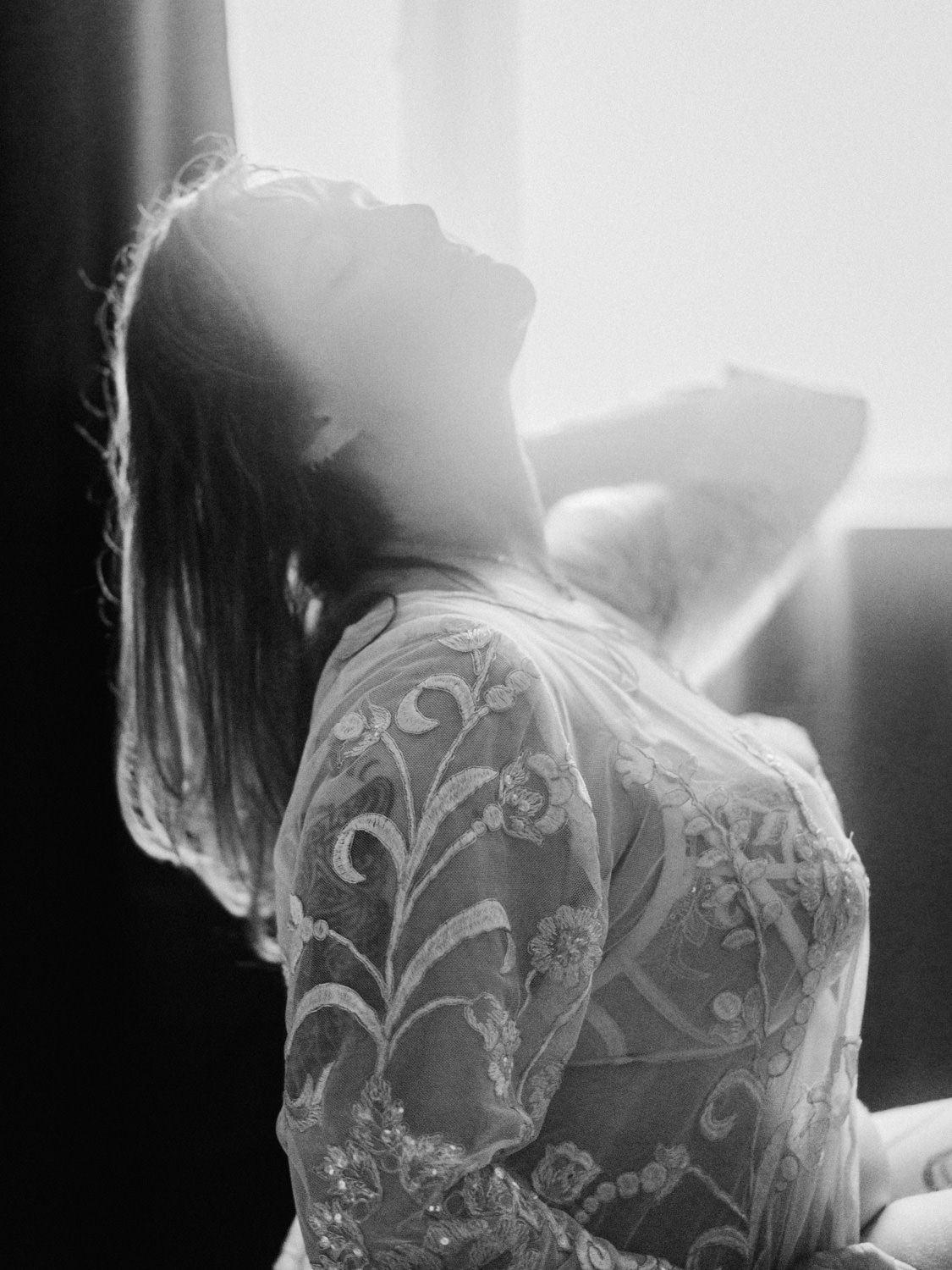 Black and white photo of a woman in lace dress tilting head back in soft backlight.