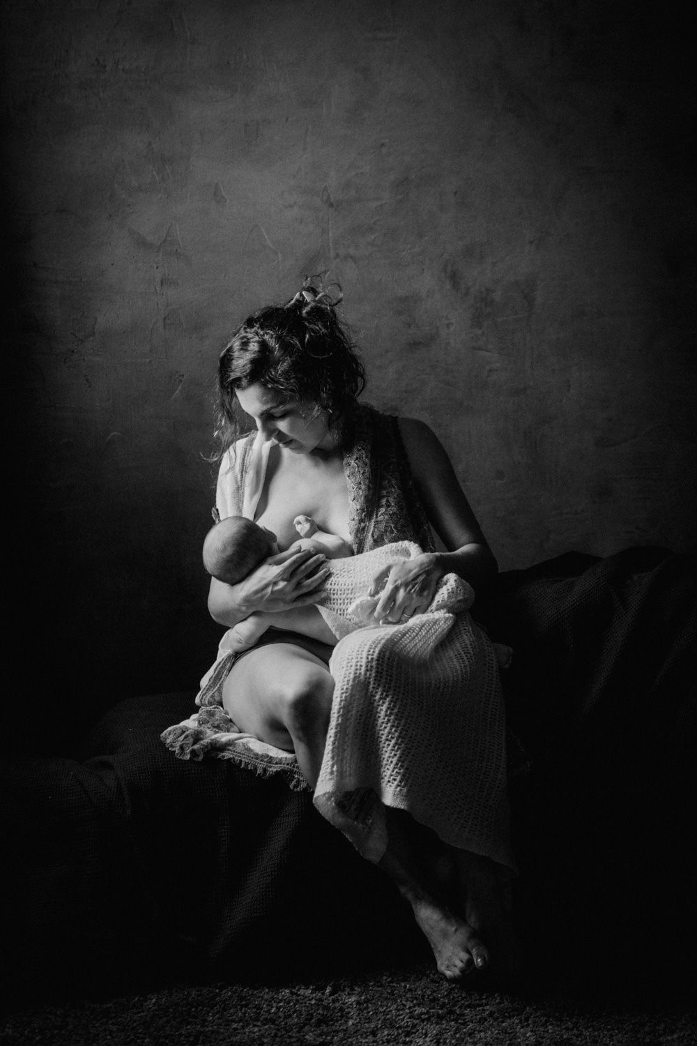 Black and white photo of a mother tenderly breastfeeding her newborn baby while seated in soft studio lighting.