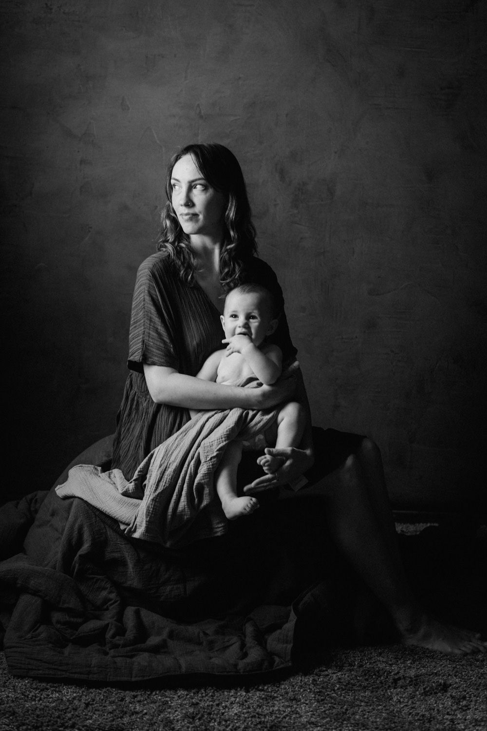 Black and white portrait of a young mother sitting and tenderly holding her smiling baby against a dark studio backdrop.
