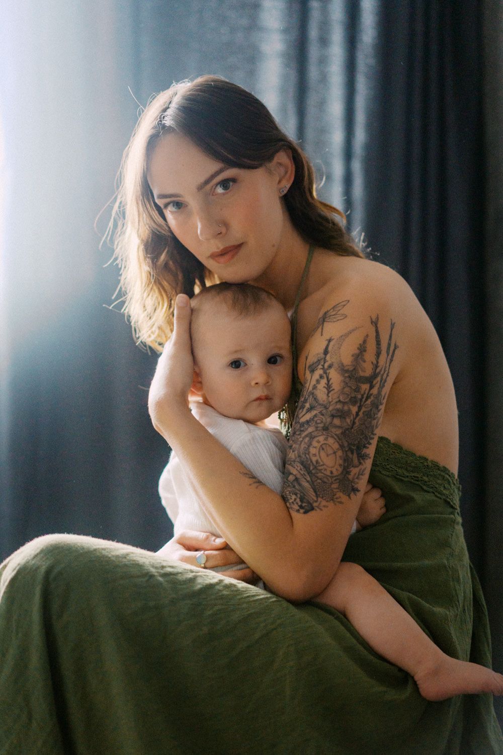 Tattooed mother tenderly holding her baby, wearing a green skirt, posed against a dark curtain backdrop.