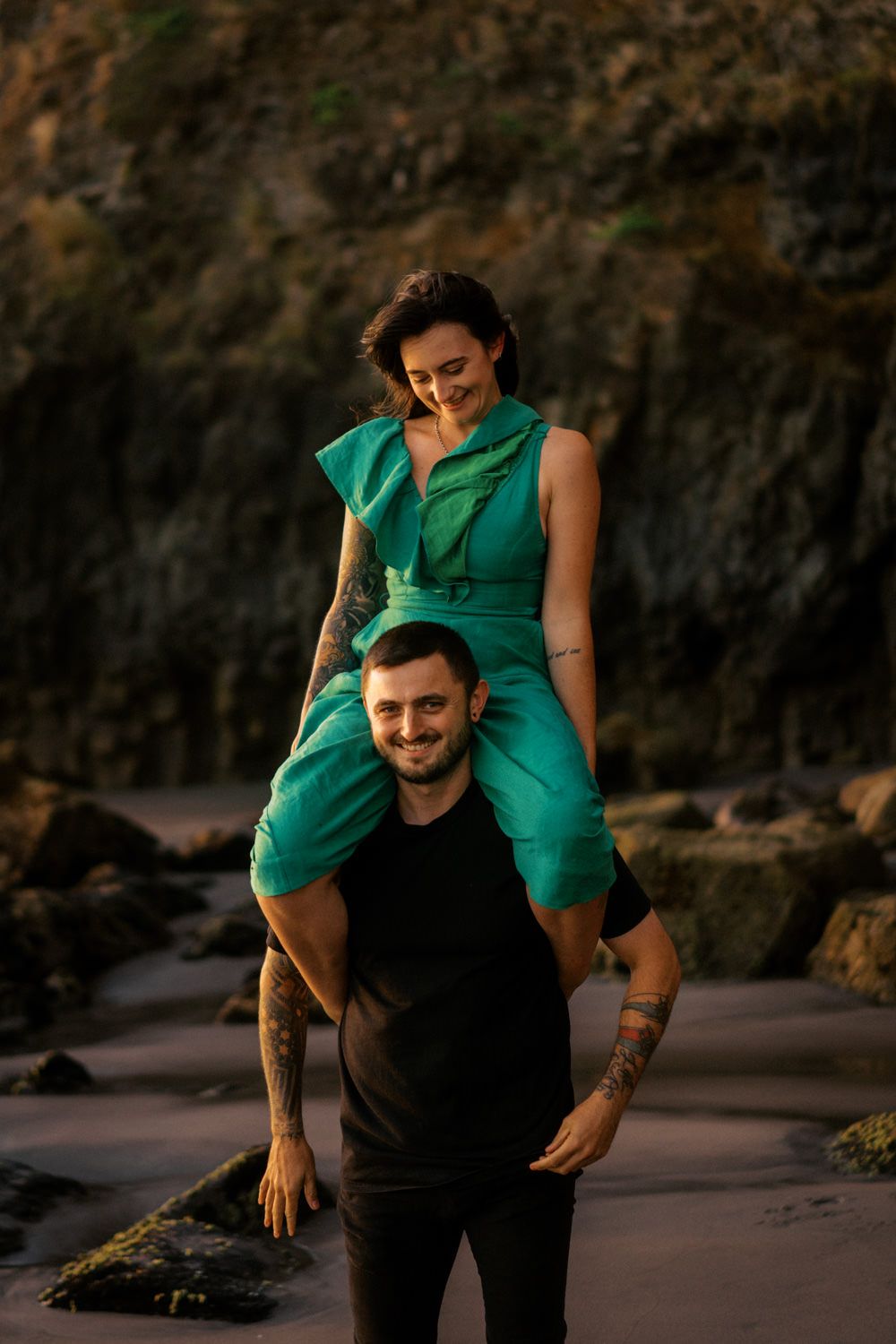 A smiling couple poses on a rocky beach, woman in teal dress sitting on mans shoulders, both with tattoos.