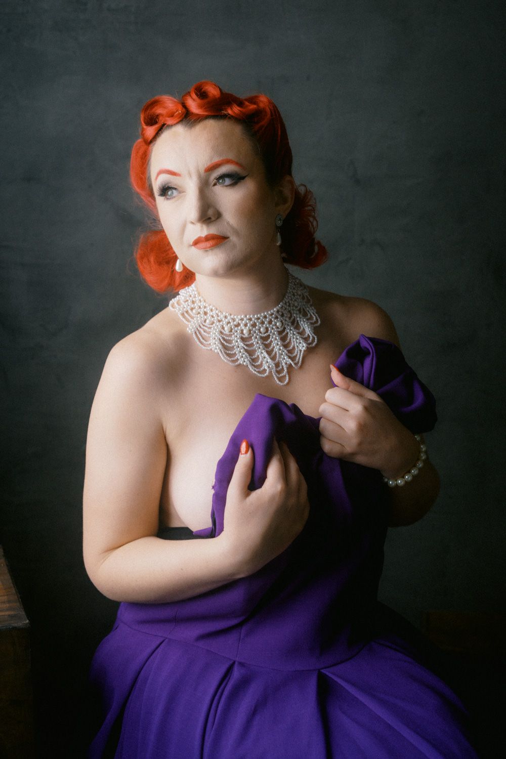 Vintage pin-up woman with red hair in purple gown, pearl necklace and bracelet, posing against dark background.
