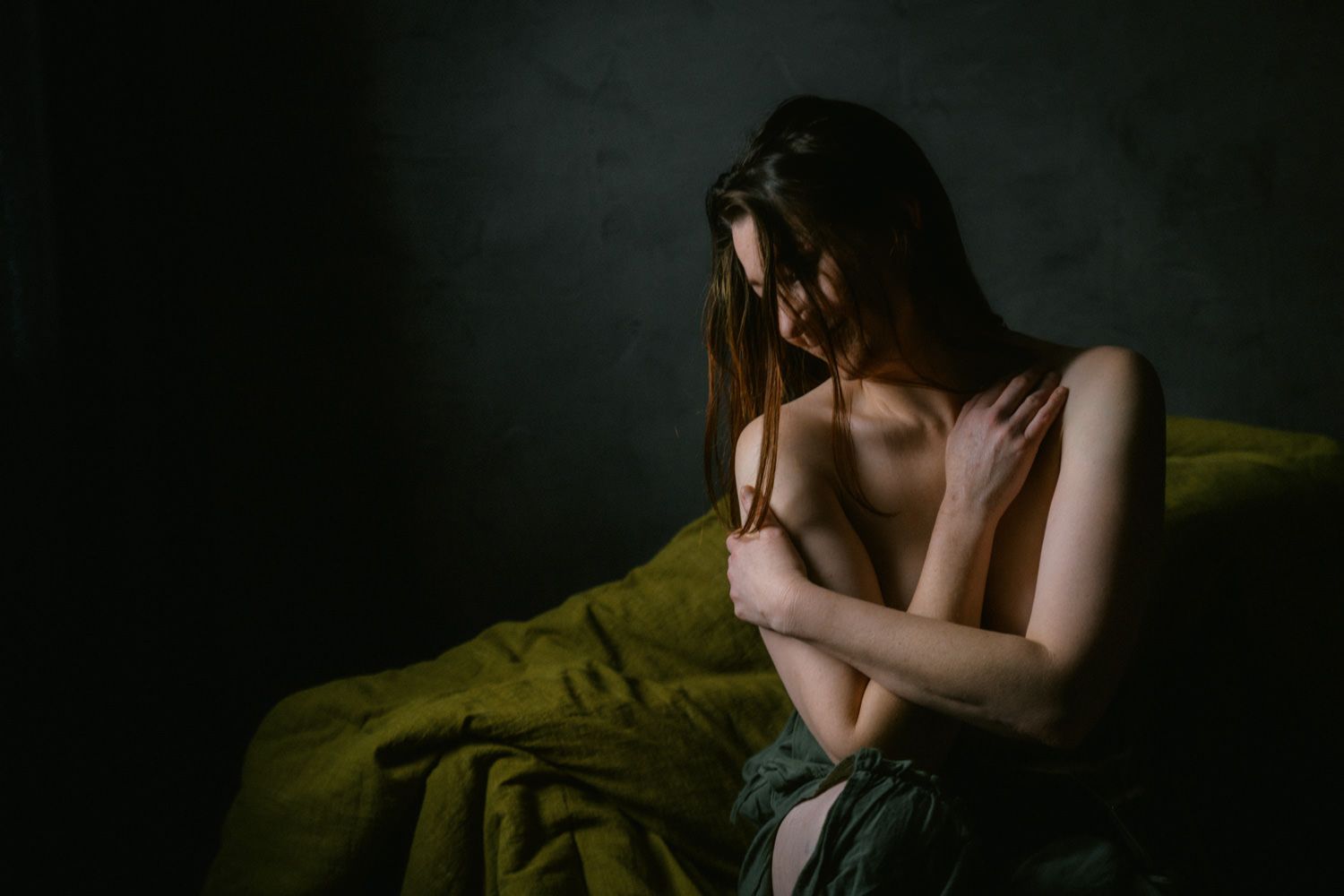 A woman sits contemplatively on a bed with green sheets in a dark, moody artistic boudoir photograph.