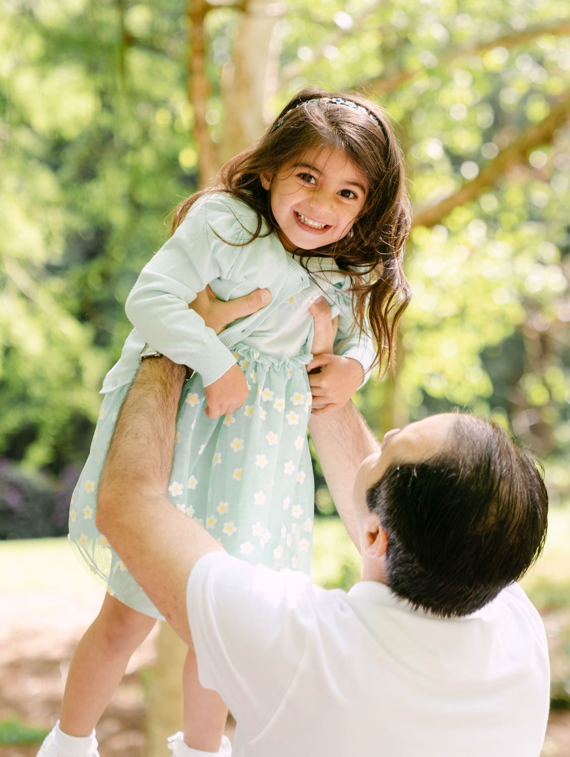 A joyful little girl in a mint floral dress being lifted by her father outdoors in a lush green park.