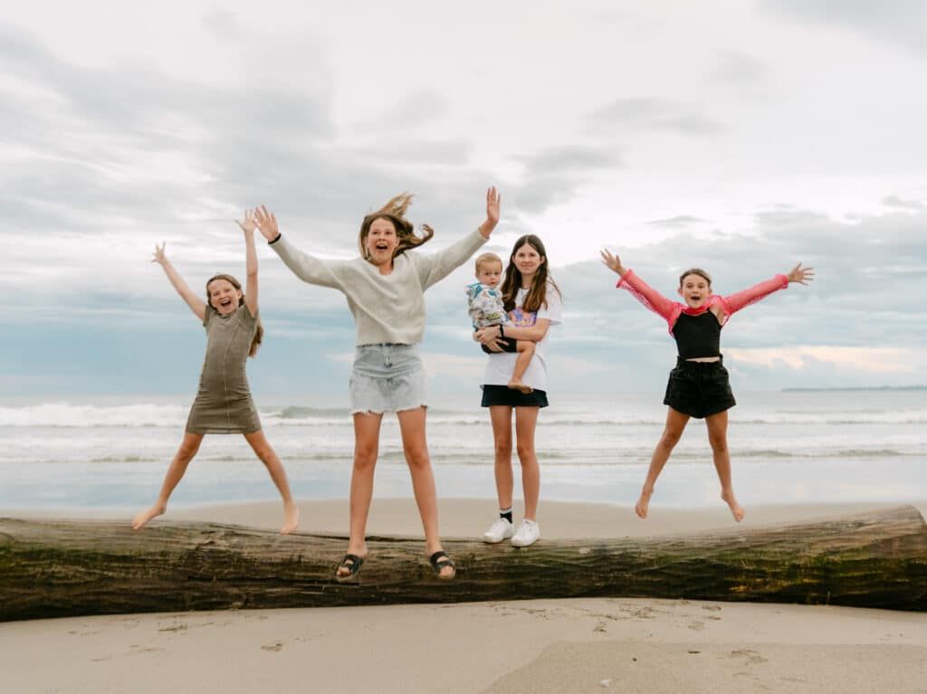Group of girls jumping joyfully on a beach log with arms raised, posing for a fun family photo by the ocean.