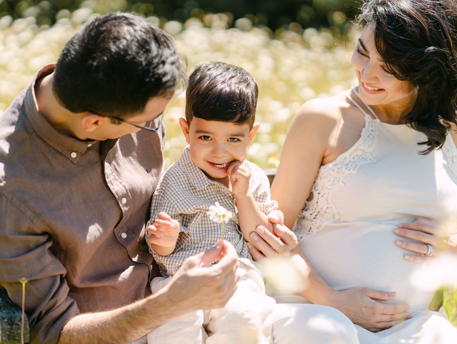 Pregnant woman and man sit outdoors with toddler boy in sunny field during maternity family photo session.