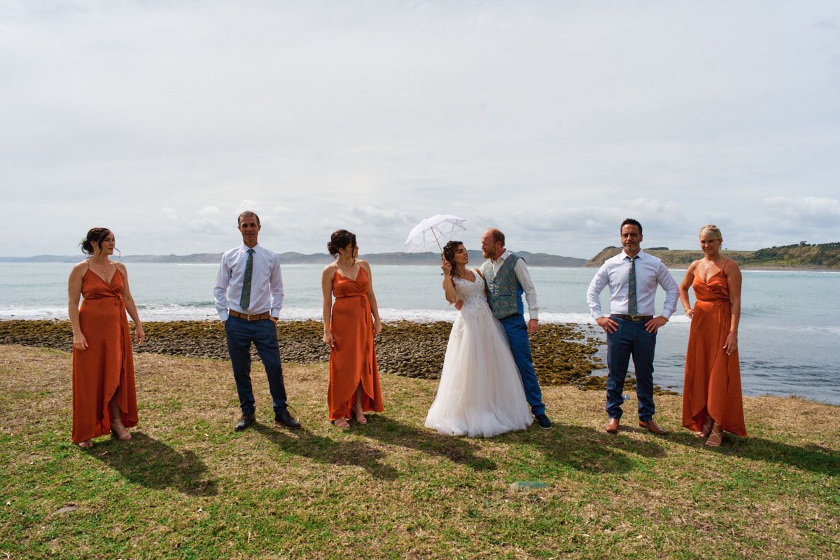 Wedding party poses outdoors near water, bride holding umbrella, bridesmaids in orange dresses, groomsmen in white shirts.
