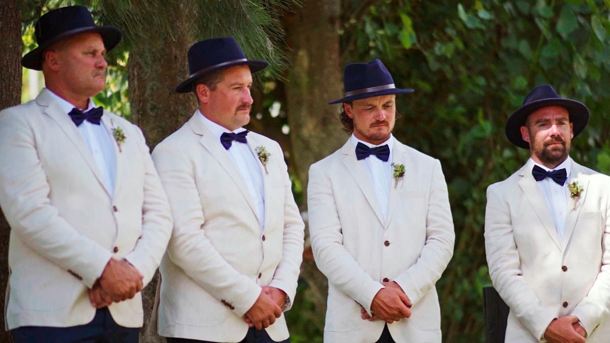 Four groomsmen in white suits, navy bow ties, and matching hats stand outdoors at a wedding ceremony.