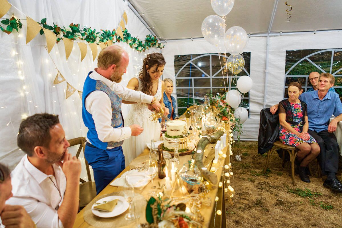 Bride and groom cutting wedding cake inside decorated marquee tent with guests seated at long table.